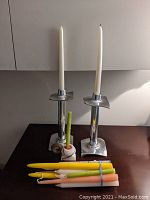 Photo of three silver tone candlestick holders with square bases and square drip trays. Candlesticks are white, pale yellow, and green. Additional loose candlesticks in yellow, white, and peach colors.