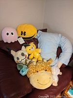 Photo showing lot of assorted plush toys arranged on a chair including a giraffe, yellow bear, pink octopus, Mimikyu, Pacman ghost, and a light blue pillow shaped like a person.