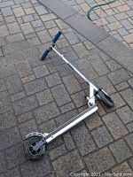 Micro kick scooter laying on pavement showing blue foam grips, silver frame, black wheels, and deck grip tape.