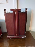 Closed view of the compact folded wooden easel showing metal clasps and carrying strap