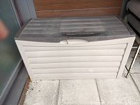 Top and front perspective of the outdoor resin deck storage box showing ribbed lid with handle and locking mechanism. Beige color with some surface dust and dirt visible.