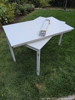 Two white Ikea Melltorp tables stacked on grass outdoors. One table lies flat on the ground while the other rests on top of it along with some metal parts on the tabletop.