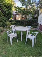White round plastic garden table with four matching plastic chairs set on green lawn under a tree, outdoor setting with houses in background.