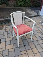 Side angled view of wooden chair with white painted frame and faded red upholstered seat cushion on outdoor patio.