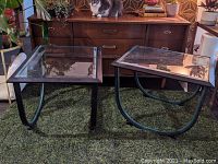 Two metal side tables with glass tops shown on green carpet in front of a wooden dresser. One table has straight legs, the other curved legs. Cat sits on dresser in background.