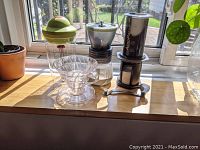 Photo showing all four items in the lot: Bodum milk frother, Aeropress coffee maker, Hario V60 dripper, and Hario Skerton grinder on a windowsill with outdoor garden background.