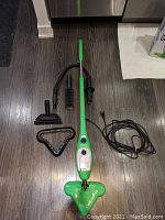 Green and white H2O Mop X5 steam mop with attached power cord and several cleaning attachments displayed on wood floor.