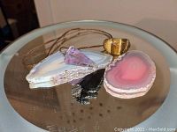 Displays pink and white geode slice, amethyst cone pendant, tiger's eye pendant, and black arrowhead together on glass surface