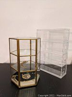 Photo showing golden framed hexagonal glass jewelry box containing gold chain bracelet and lion pendant, next to a clear four-drawer acrylic Muji glasses case.