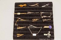 Photo showing all 14 assorted tie clips arranged on a black display pad, showcasing variety in metal tones, chain details, and shapes including a sterling silver marked clip with charm.