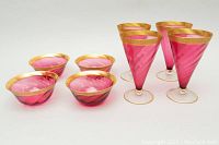 Four cranberry footed glasses and four cranberry dessert bowls all featuring gold rims, shown on a white background.