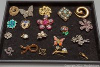 Full view of all 20 brooches arranged on a black felt background showing various styles including floral, butterfly, and abstract designs with some stones missing and wear visible.