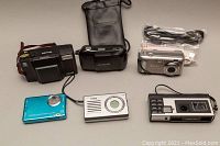 Six assorted electronic devices including cameras and MP3 player displayed on flat surface with cables and carrying pouch