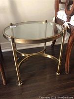 Round brass and glass side table positioned near wooden furniture. The table has a clear glass top with a circular brass frame and three brass legs.
