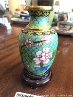 Front view of the cloisonne vase showing floral motifs and gold accents on turquoise background, placed on wooden stand.