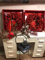 Two boxed large red floral wreaths, silver candle holder, plaid basket with red bow, Halloween-themed metal lantern on a filing cabinet.