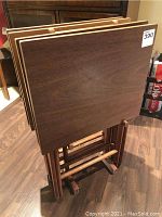 Four mid century modern style wooden tray tables folded and standing upright in their matching wooden holder. Each table has a smooth wood grain veneer top with light trim edges, and cylindrical folding legs connected by metal hinges.