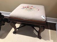 Side angled view of the vintage needlepoint stool showing curved wooden legs and floral embroidery on the padded seat.