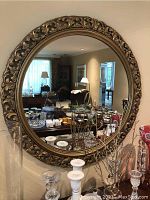 Full view of the round mirror mounted on a wall, showing its large size and the detailed gold frame, reflective surface displays furniture in background.