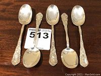 Five Marsh & Hoffman sterling silver spoons laid out on a wooden table, showing front view with ornate handles and bowl shape.