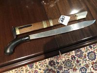 Tribal blade with wooden sheath fully displayed on wooden surface, showing curved dark handle and metal fittings.