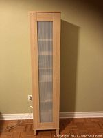 Front view of tall corner cabinet showing light wood and frosted glass door panel.
