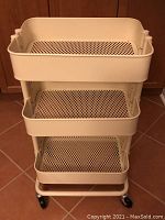 Front view of the IKEA white metal storage rack showing three mesh shelves and caster wheels on a tiled floor.