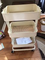 Full front view showing three white metal mesh shelves stacked vertically with adjustable supports and caster wheels at bottom.