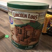Closed green metal tin container with Lincoln Logs branding and 100th Anniversary label, showing log cabin images.
