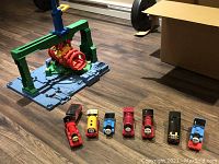 Six different plastic Thomas trains positioned in front of a green and blue crane playset on wood floor.