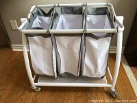 Front view of the three-section laundry basket showing white metal frame, gray and white fabric baskets, and caster wheels.