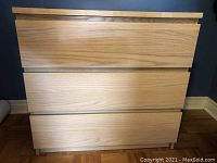 Front view of wooden chest with three wide drawers and flat, handle-less fronts in light oak finish against a blue wall.
