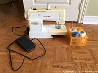 Front view of Singer sewing machine with foot pedal cord and orange basket containing threads and oil bottle.