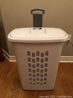 Front view of white laundry basket with ventilation holes and gray handle extended, shown upright on hardwood floor.