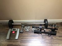 Full view of various gym items including barbell with plates, dumbbell, two loose 10 lb plates, fitness scale, ab roller, and hand grips on wooden floor against beige wall.