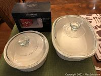 Two white ceramic CorningWare bakeware dishes with glass lids and boxed bone china mugs displayed on table