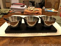 Set of three hammered metal bowls with small black spoons sitting in a rectangular dark wood tray sitting on a white cloth on a table with other household items in background.