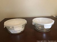 Two round clear glass food storage containers side by side on a wooden surface, each with white plastic locking lids secured.