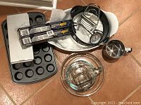 Photo shows a variety of bakeware including Pyrex measuring cups, muffin tins, glass pie dish, metal cooling rack with thermometer, and kitchen tools such as egg slicer and flour sifter.