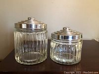 Two round glass containers with vertical ridged design and silver-colored metal lids with concentric rings and knob handles.