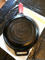 Bottom view of Lodge cast-iron skillet showing cast iron material and model number P12S, with attached red silicone handle cover, photographed on paper towel and black plastic table cover.
