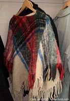 Multicolor plaid poncho draped on hanger showing red, green, blue and cream panels with fringe trim