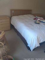 View of entire bed with light solid wood headboard and white bedding, metal frame underneath, nightstand next to bed.