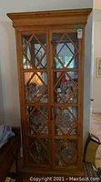 Front view of tall wood and glass display cabinet showing double glass doors with diamond mullion design, lock with key, and visible interior glass shelves with contents inside (not included)