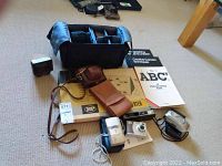 Wide view of the lot showing the Optex camera bag with padded compartments, multiple cameras, lenses, flash, and slide viewers laid out on floor.