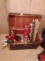Photo of open vintage chest filled with Christmas decorations including fabric reindeer, nutcrackers, wrapping paper, ornaments, ribbons, and bows.