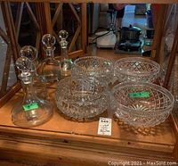 Full view of two cut crystal salad bowls with intricate patterns and two clear glass decanters with stoppers, displayed on wood shelf.