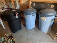 Photo showing three plastic garbage bins: one black with wheels and two blue without wheels placed on concrete floor.