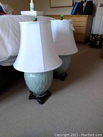 Two matching ceramic lamps with white shades placed on carpeted floor beside bed. Lamps have pale green ceramic bases with leaf pattern and dark wooden bases.