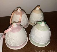 Four Lladro Christmas bell ornaments in white porcelain with colored ribbons and bases, displayed together.